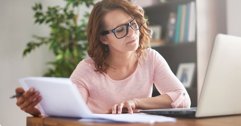 a woman looking to her laptop while holding a bunch of paper